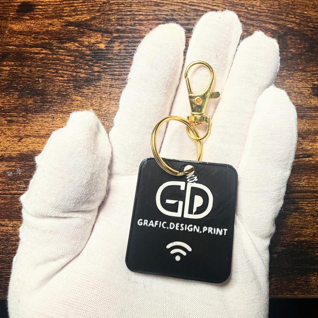 Black keychain with 'GD Grafic Design Print' logo held in a white gloved hand on a wooden surface.