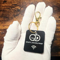 Black keychain with 'GD Grafic Design Print' logo held in a white gloved hand on a wooden surface.
